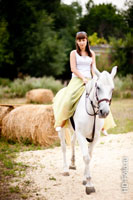 Фото девушки в белом топе и длинной светлой юбке верхом на белой лошади
