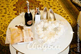 Фото белых букв «Свадьба» на белом столе, чулок и туфель невесты, свадебных колец, бутылки шампанского с бокалами