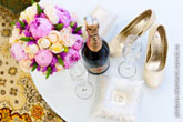 Фото свадебного натюрморта сверху на белом столе: красивый букет невесты, бутылка шампанского с бокалами, обручальные кольца на подушечке, туфли невесты