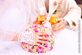 Фото руки невесты с обручальным кольцом, лежащей на свадебном букете, бокалы с шампанским в руках на дальнем плане